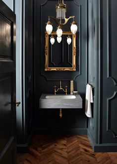 a bathroom with black walls and wood floors, gold framed mirror on the wall, marble sink