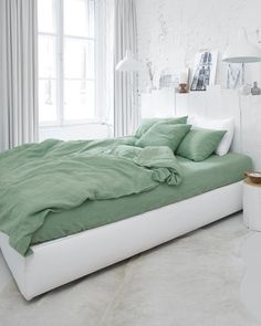 a bed with green sheets and pillows in a white room next to a large window