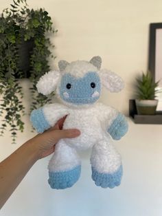 a hand holding a small white and blue stuffed animal next to a potted plant