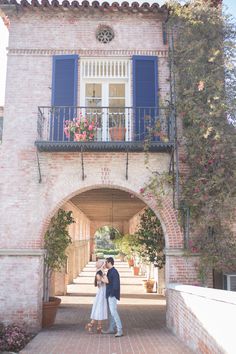 an engaged couple standing in front of a building with blue shutters and flowers on the balconies