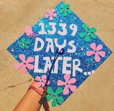 a hand holding a blue graduation cap with flowers on it that says, 123 days later