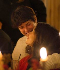 two people sitting at a table with candles in front of them and one person kissing the other