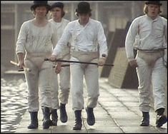 four men in white suits and hats walking down the street with one holding a stick