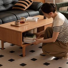 a woman kneeling down next to a coffee table with books on it and a black couch in the background