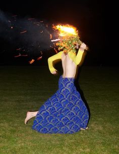 a woman sitting on the ground with fire in her hair and holding something up to her head