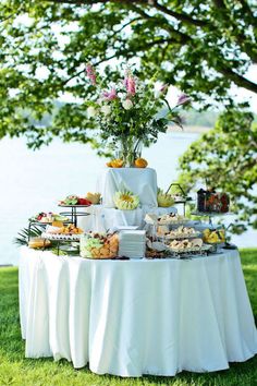 a table topped with lots of food on top of a lush green field next to water