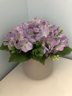 a white vase filled with purple flowers on top of a table next to a wall