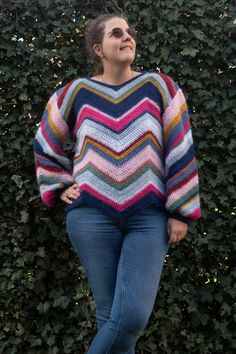 a woman standing in front of a hedge wearing a multicolored sweater and jeans