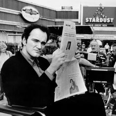 a man sitting in a chair reading a newspaper while people stand around him and look on