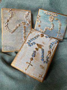 two wooden plaques with beads on them sitting on a blue cloth covered surface, one has a cross and the other is a rosary