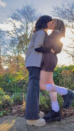 two people standing next to each other in front of trees and bushes, one kissing the other