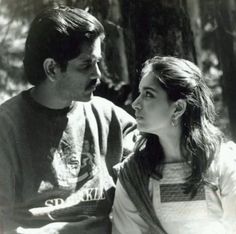 a man and woman sitting next to each other in the woods looking at each other