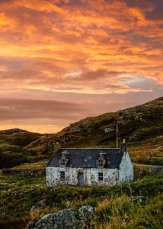 an old house sitting on top of a lush green hillside under a colorful sky with clouds