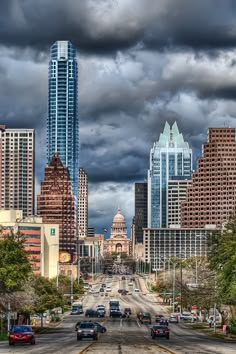 cars driving down the road in front of tall buildings under a cloudy sky with dark clouds