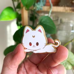 a hand holding a white cat brooch in front of a green potted plant