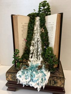 an open book with trees and waterfall in the middle, on a table next to books