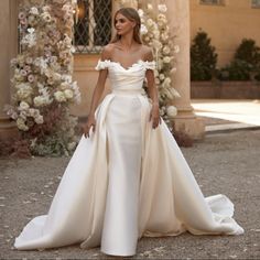 a woman standing in front of a building wearing a wedding dress with an off the shoulder neckline