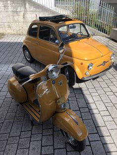 an orange scooter parked next to a yellow car on a brick sidewalk in front of a fence