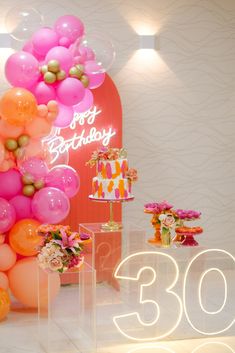 the birthday cake is surrounded by balloons and flowers on display in front of a neon sign