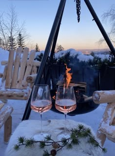 two glasses of wine sitting on top of a table covered in snow next to a fire pit