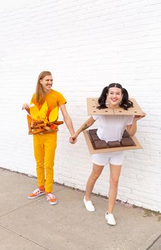 two women holding hands and standing in front of a white brick wall with an open cardboard box on their head