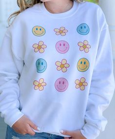 a woman wearing a white sweatshirt with smiley faces and flowers on the front, standing in front of a window