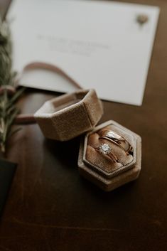 two wedding rings sitting on top of a wooden table next to a ring box and some greenery