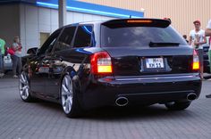 the back end of a black car parked in front of a building with people standing around