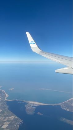 the wing of an airplane flying over some water and land in the distance with blue sky