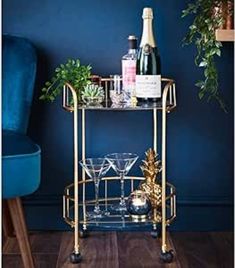 a gold bar cart with drinks on it in front of a blue chair and potted plant