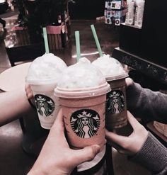 three starbucks drinks are being held up by two people at a table in a coffee shop