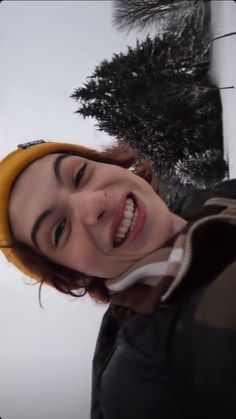 a woman smiling and wearing a yellow beanie on top of her head with trees in the background