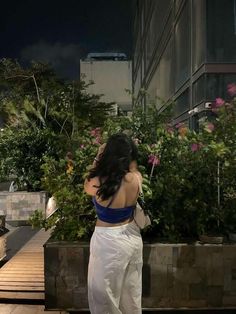 a woman in white pants and blue top standing next to some plants on a sidewalk