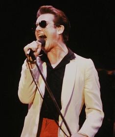 a man singing into a microphone while wearing red shorts and a white blazer on stage