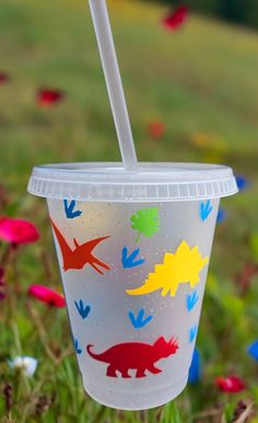 a plastic cup with a straw in it on the grass and flowers behind it is an image of dinosaurs