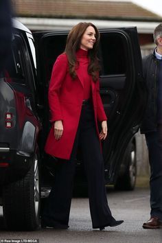 a woman in a red coat and black pants walking out of a car with another man standing next to her