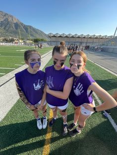 three girls in purple shirts and white shorts standing on a field with their hands painted