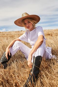 a woman sitting in the middle of a field wearing a cowboy hat and white pants