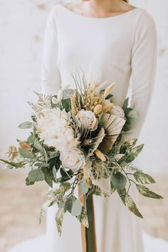 a bridal holding a bouquet of flowers and greenery in her hands, wearing a white long sleeved dress