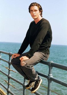 a man sitting on top of a metal railing next to the ocean and looking at the camera