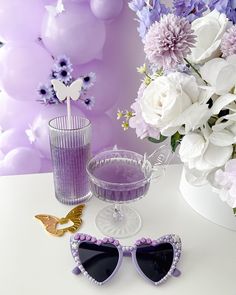 purple and white flowers are in the vase next to two sunglasses, one with butterfly shaped frames