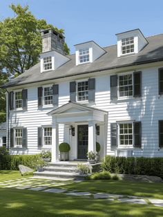 a large white house with black shutters on the front and side windows, sitting in a lush green yard