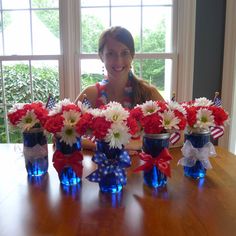 a woman sitting at a table with four vases filled with red, white and blue flowers