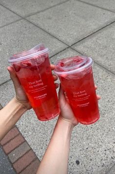 two people holding up cups with drinks in them on the sidewalk, one is red