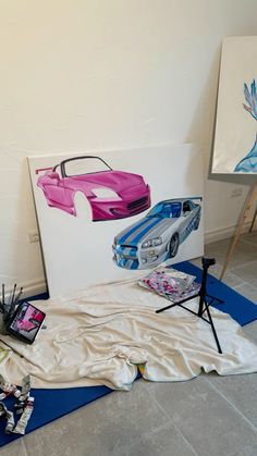 some art work is being displayed in front of two paintings on easels, one with pink and blue cars painted on it