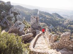 two people walking up the side of a stone wall next to trees and mountains in the background