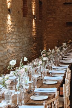 a long wooden table with white plates and flowers in vases on the tables set for dinner