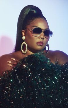 a woman wearing sunglasses and a black dress with sequins on the shoulders, standing in front of a pink background