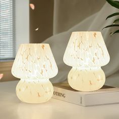 two white lamps sitting on top of a table next to a book and potted plant