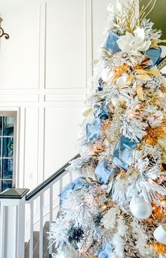 a white christmas tree with blue and gold ornaments on it's branches in front of a staircase